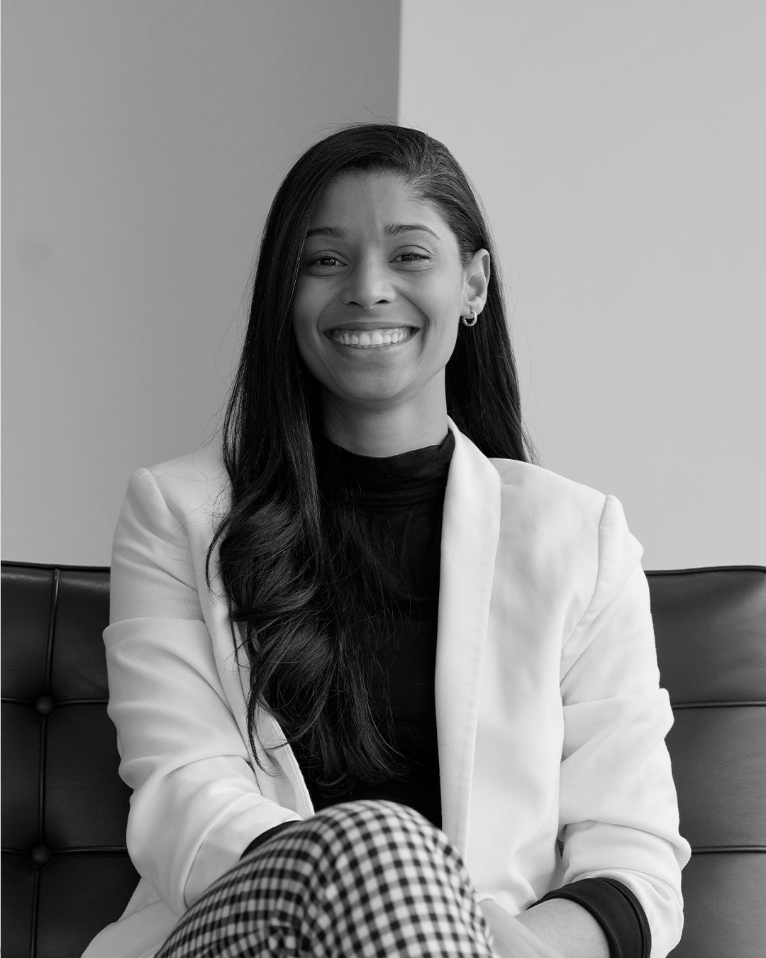 A smiling woman with long dark hair, wearing a white blazer over a black top, sitting on a black cushioned bench in front of a plain wall.