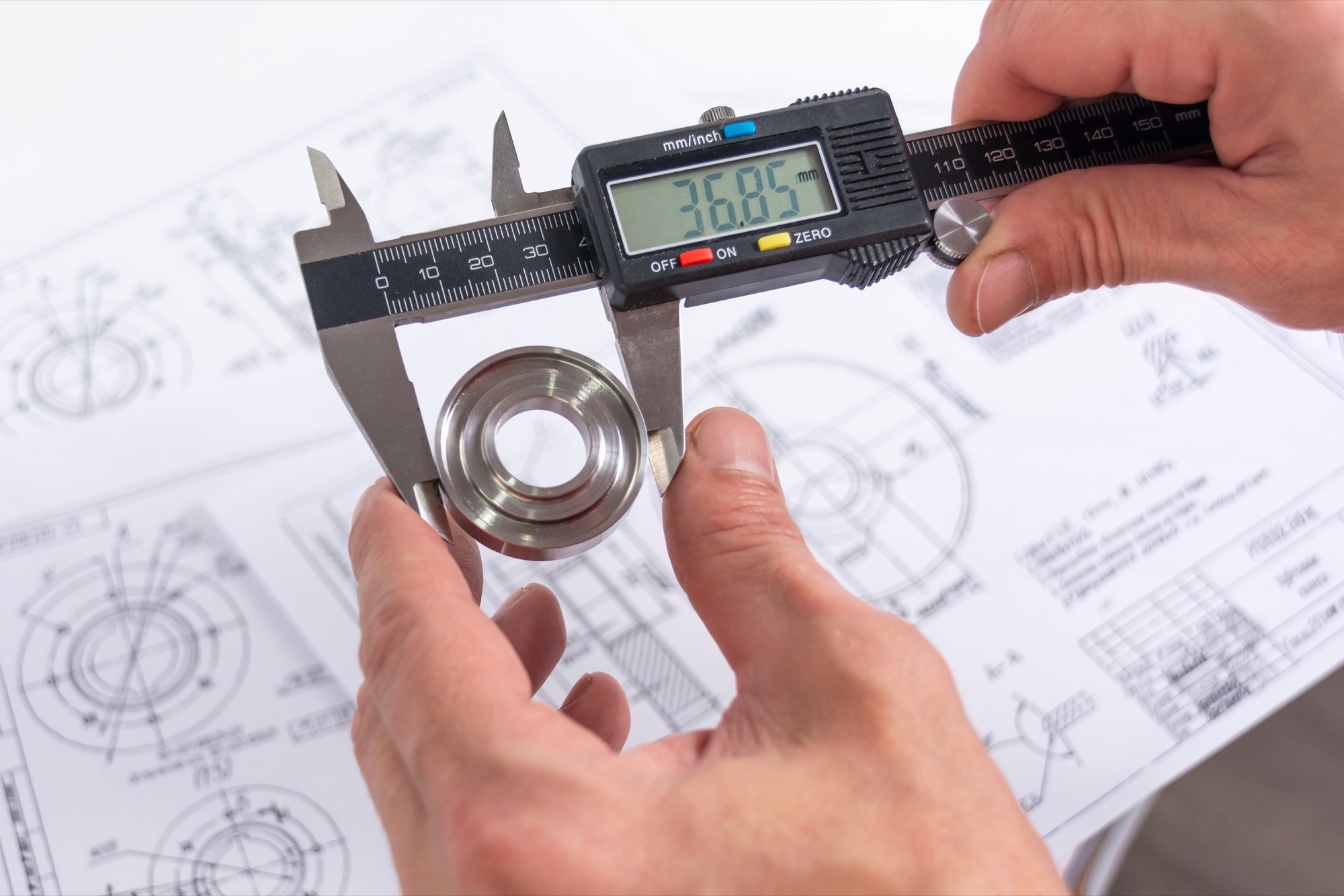 Person using a digital caliper to measure the diameter of a metal bearing, with engineering drawings in the background.