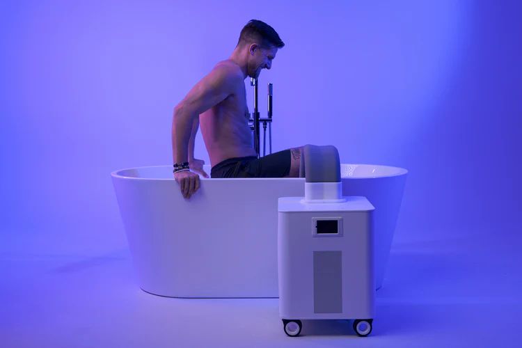 Home Plunge cold plunge A man is sitting in a modern white bathtub with a faucet and a digital device in a blue-lit room.