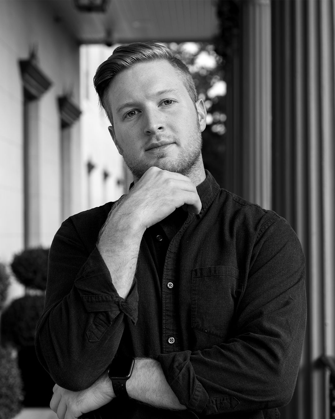 A black and white portrait of a young man with short, styled hair, wearing a dark shirt, standing outdoors next to a building with windows and a metal wall, with trees in the background. He is looking at the camera with a thoughtful expression, resting his chin on his hand.