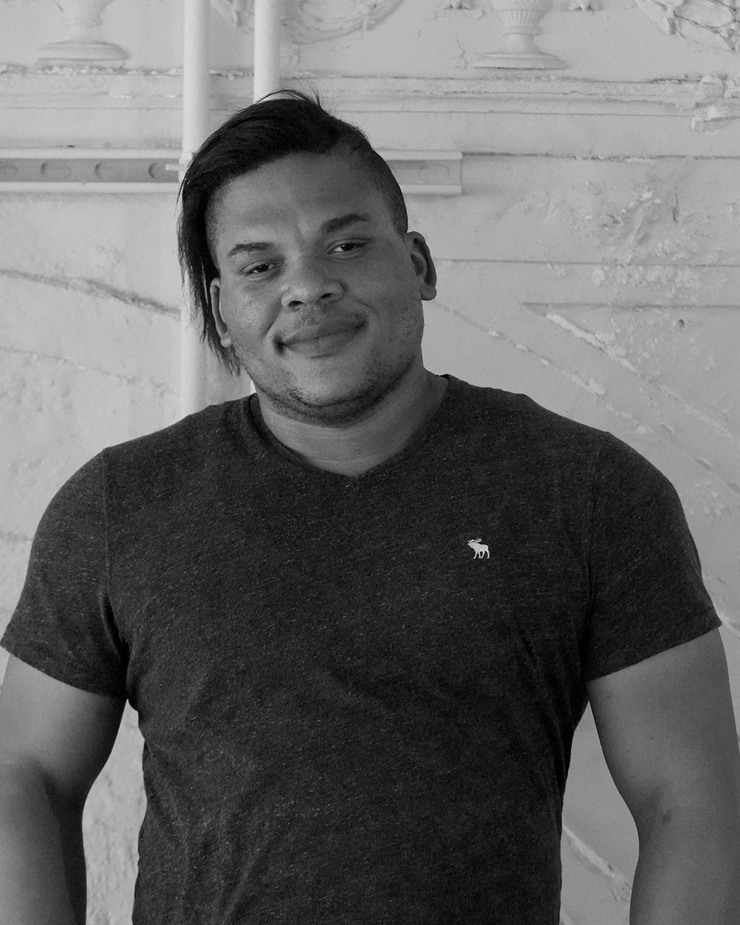 A black and white photograph of a man with a side-shaved hairstyle, wearing a dark T-shirt with a small logo on the chest, smiling slightly, standing against a textured wall with visible pipes.