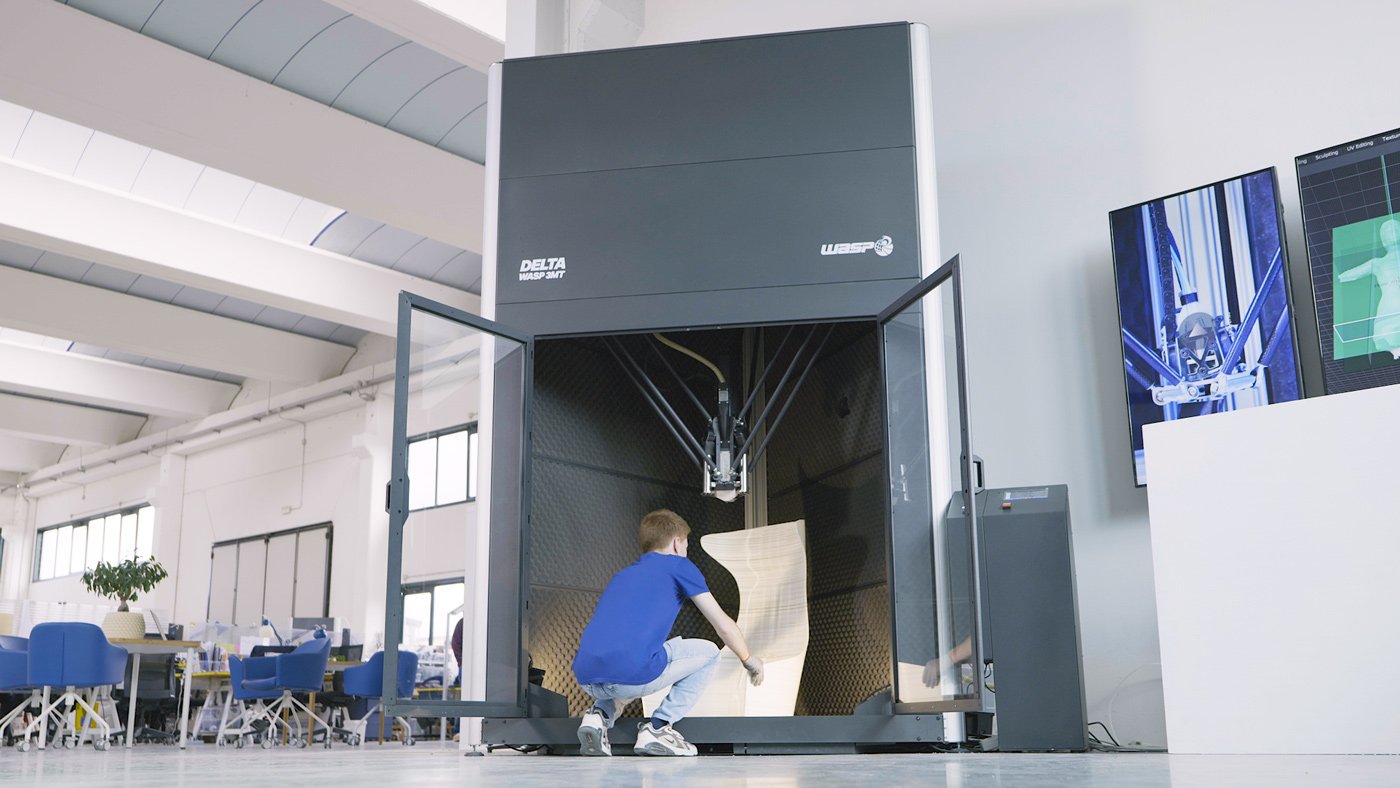 A person is operating a large 3D printer in a spacious industrial or research facility, with two digital screens displaying 3D models or print data nearby.