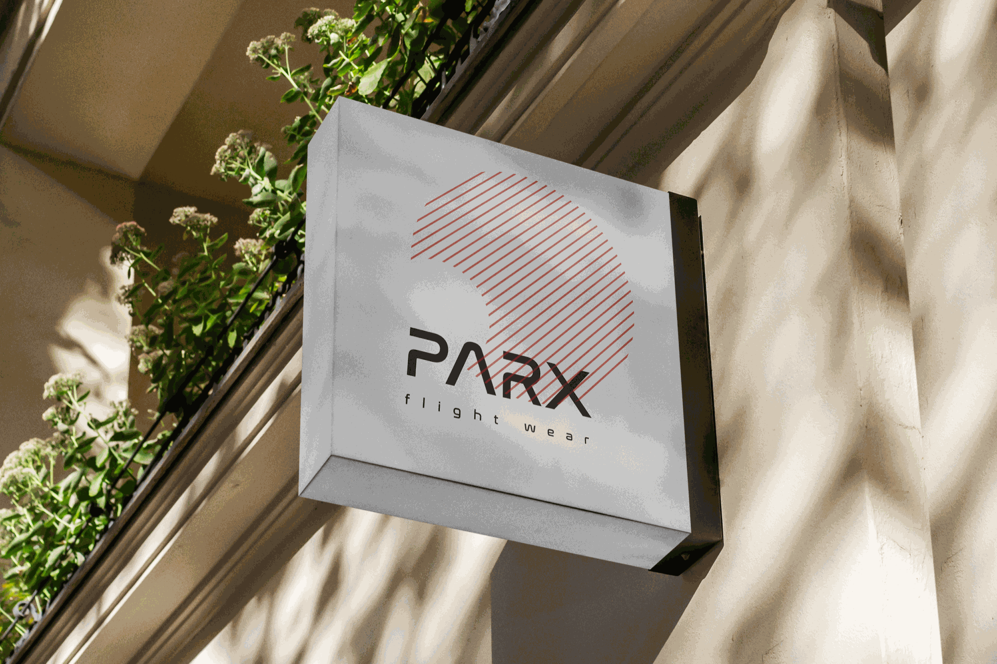 Street view of a square sign for Parax Flight Wear attached to a building with a balcony and green plants, with shadows cast on the wall.