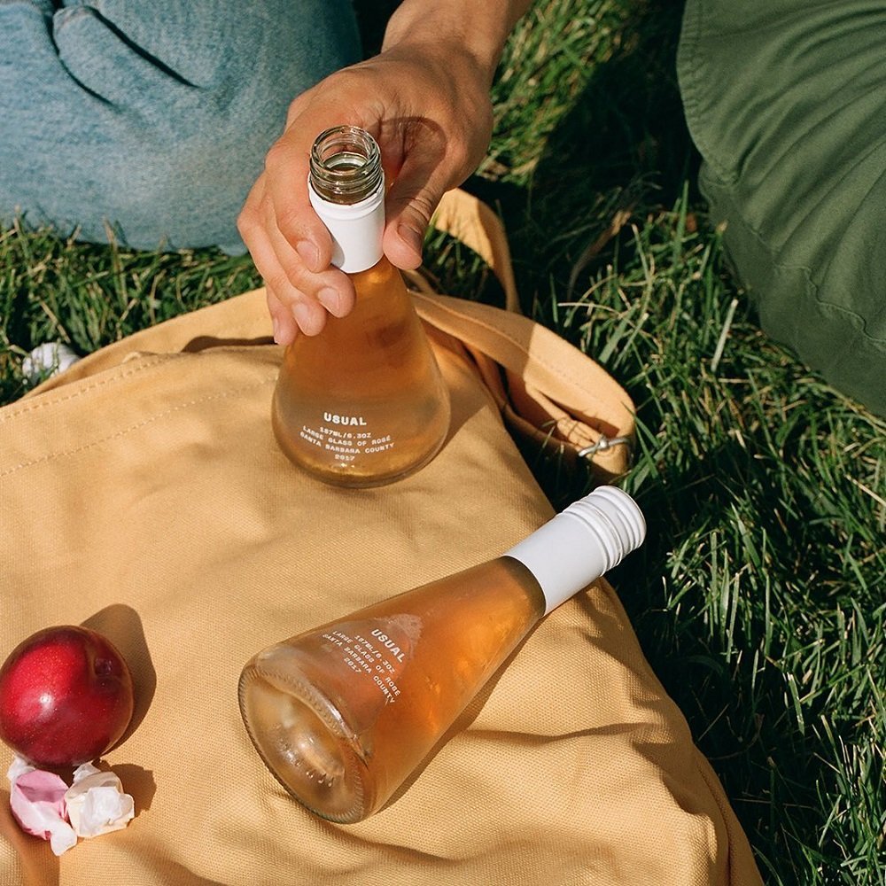 Close-up of someone opening a bottle of rosé wine on a picnic blanket outdoors. Two bottles of rosé wine, a red apple, and a crumpled paper napkin are on the blanket. Green grass surrounds the scene.