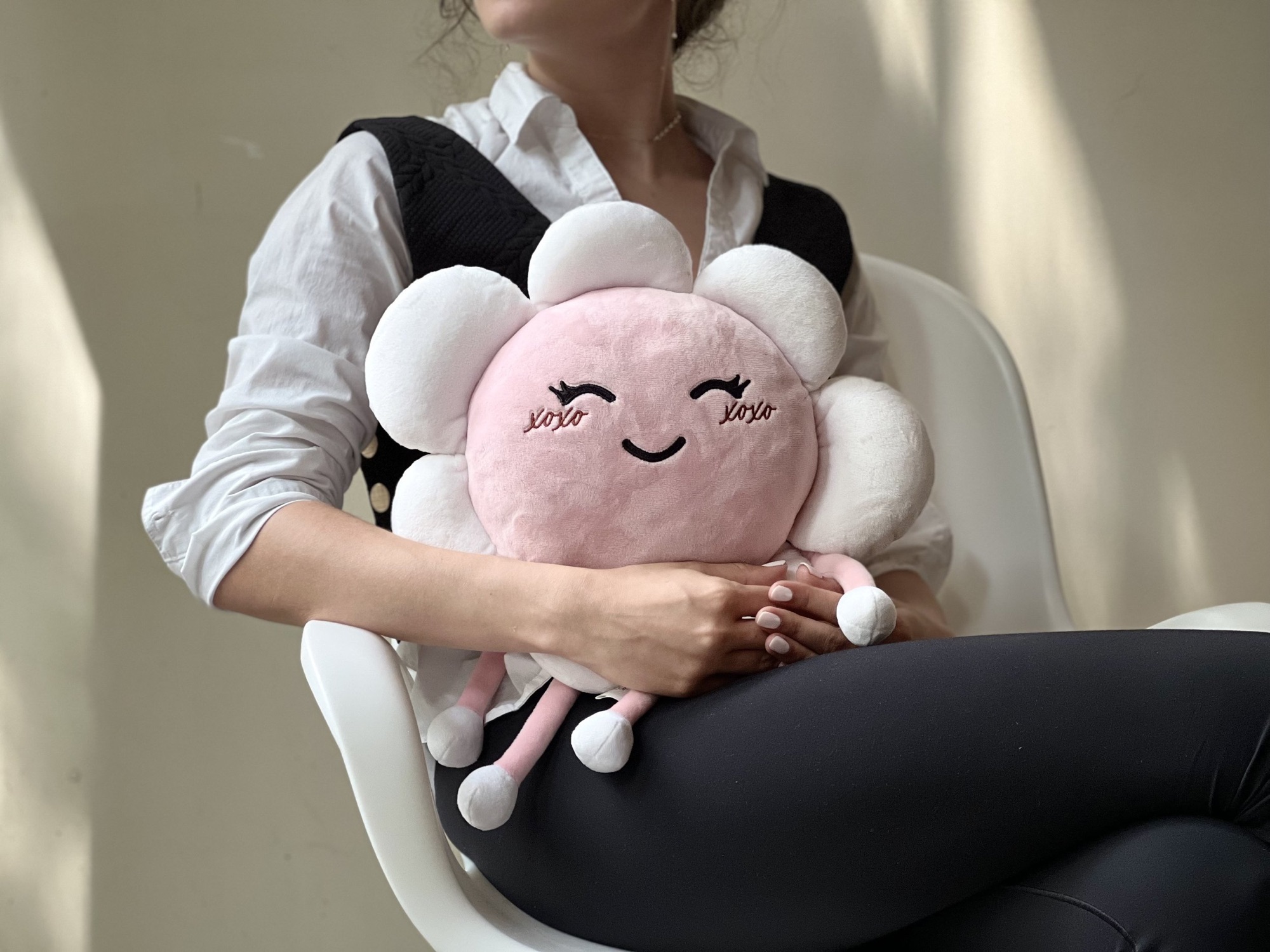 A woman sitting on a white chair holding a plush toy. The plush toy is a pink and white smiling cloud with closed eyes and little pink arms and legs.