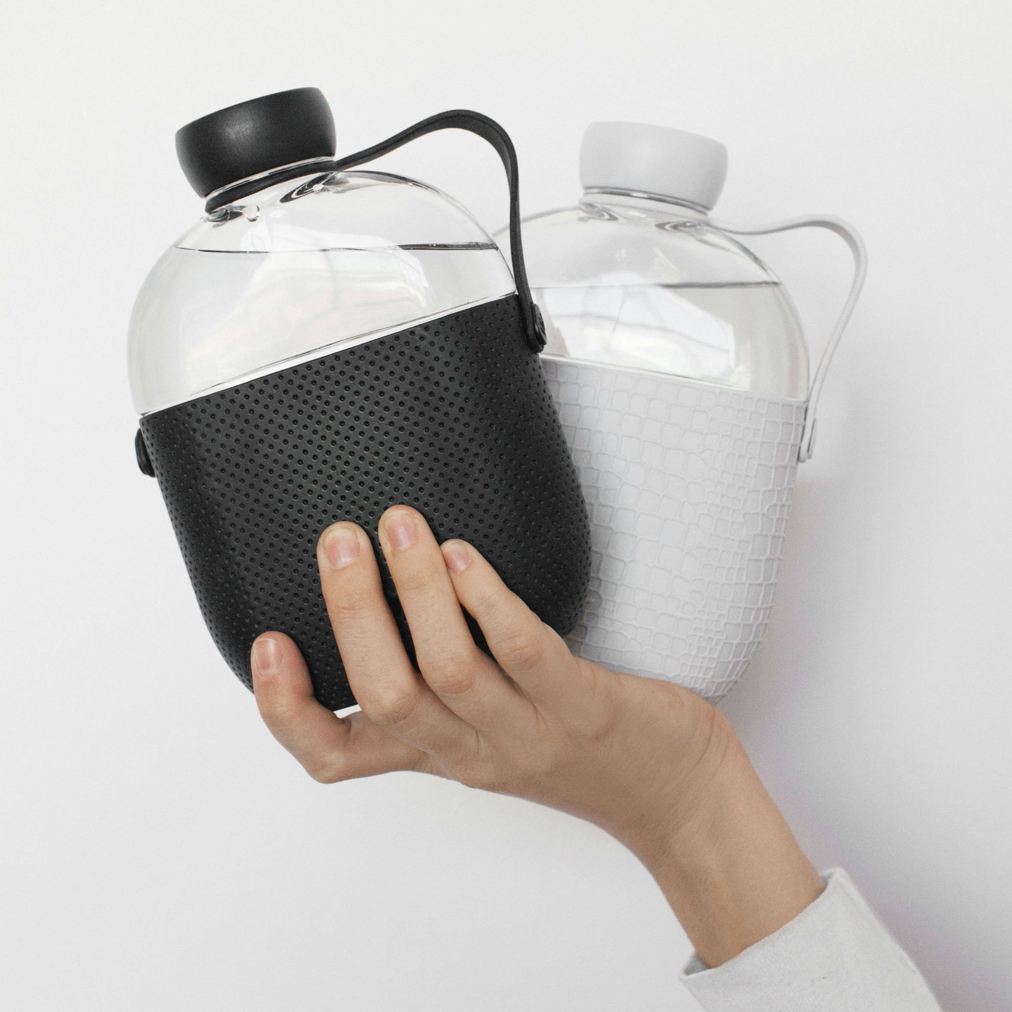 A hand holding a glass water bottle with a black textured silicone sleeve, and another similar bottle with a white sleeve in the background.