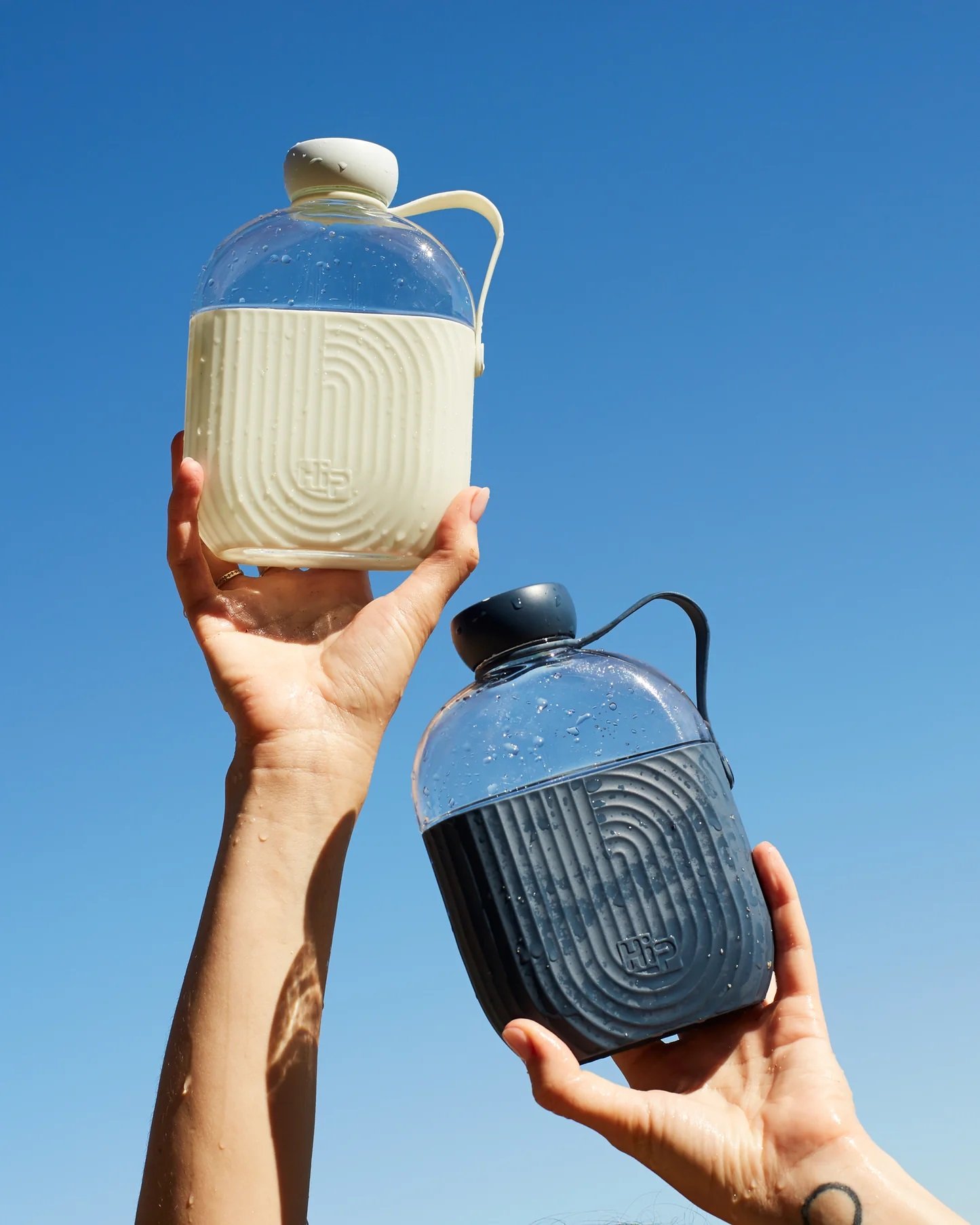Two hands holding up water bottles against a clear blue sky, one bottle is white and the other is black, both feature ridged design and are partially filled with water.
