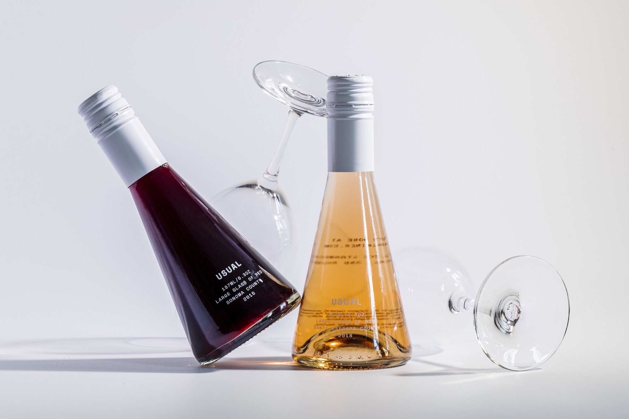 Two empty Usual single-glass wine bottles with wine glasses on a white surface
