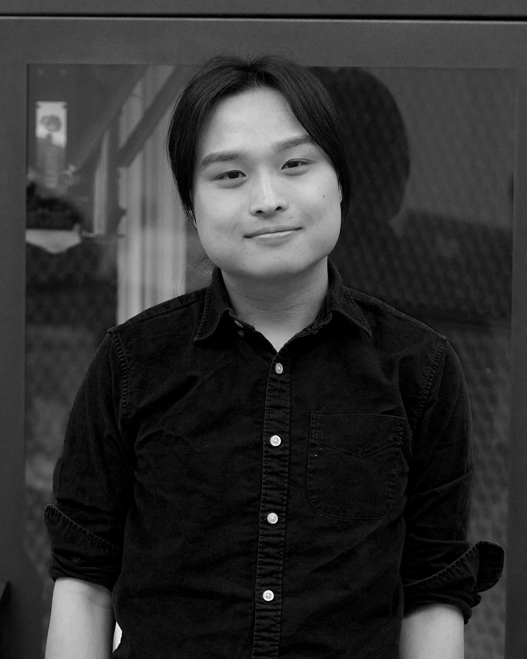 Black and white portrait of a young person with straight, dark hair, wearing a button-up shirt, standing indoors in front of a window.