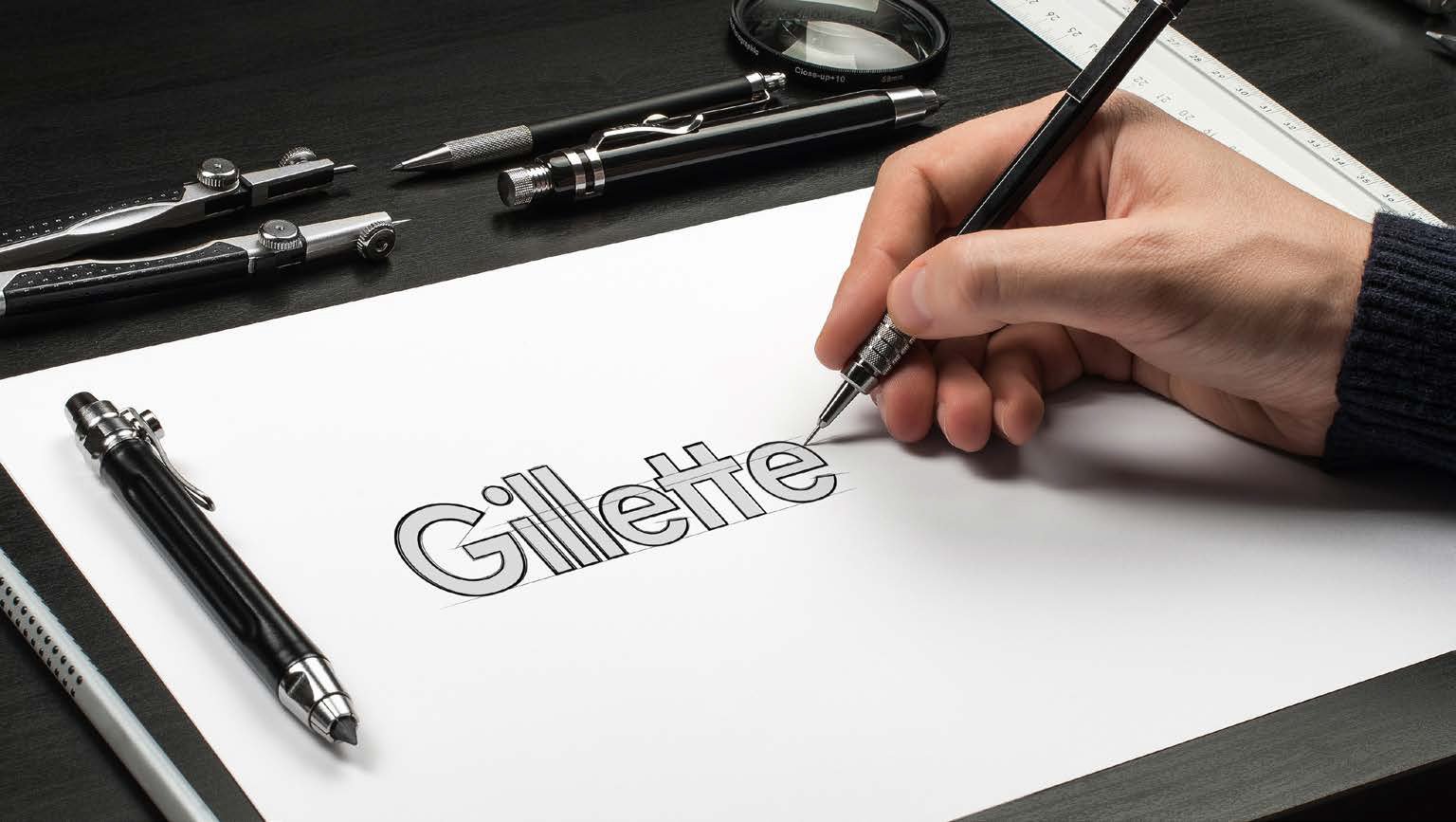 Designer sketching the Gillette logo by hand on paper with drafting tools in the background.