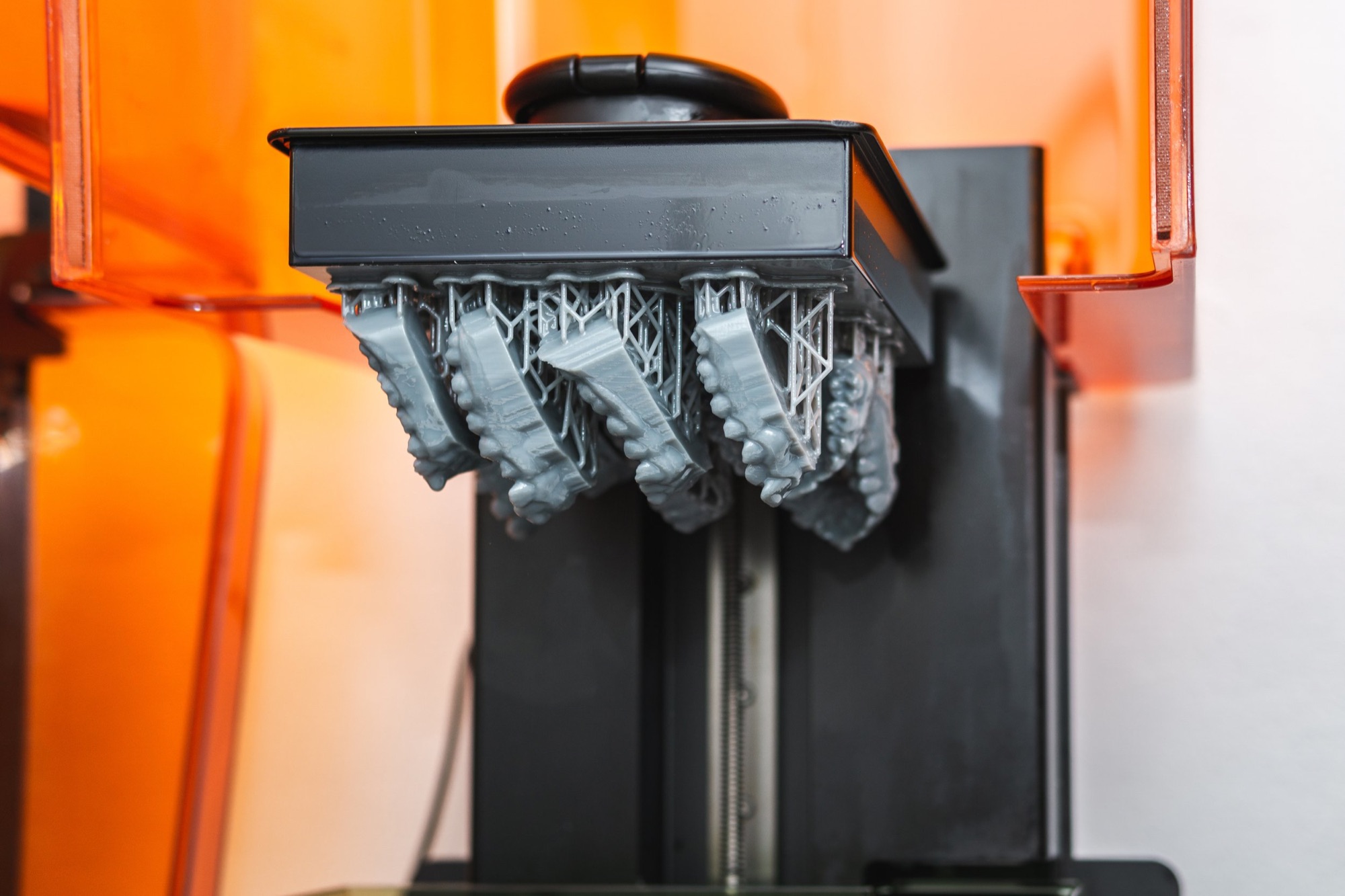 A 3D printer creating a gray object with intricate lattice structures, with orange and black parts visible in the machine's frame.