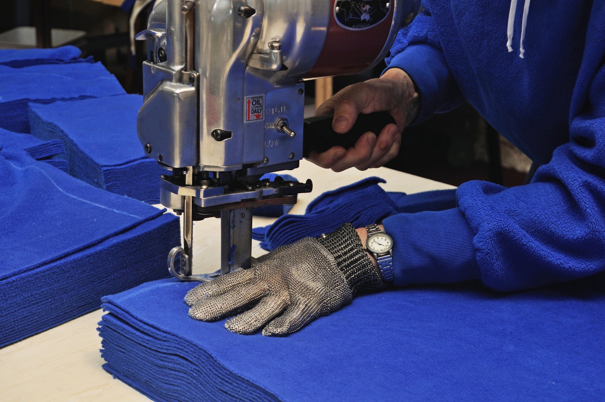 Person using a fabric cutting machine to cut blue cloth, with blue fabric stacks nearby, wearing a silver wristwatch and gray glove.