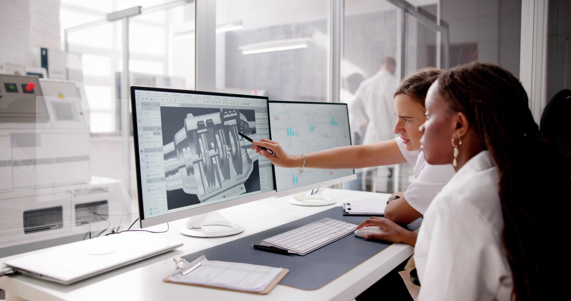 Two women analyzing a 3D CAD design on computer monitors in an office setting, with one woman pointing at the screen.