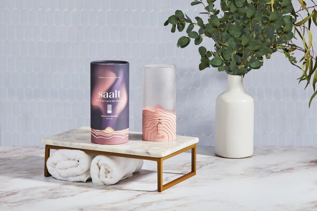 Two cylindrical containers with the brand name 'saalt' on a small marble table, next to a white vase with green leafy branches, and two rolled white towels underneath the table.