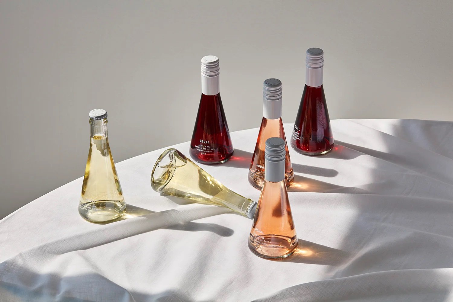 Six glass bottles of different colors and shapes, some upright and some toppled, sitting on a white cloth-covered surface with a plain light background.