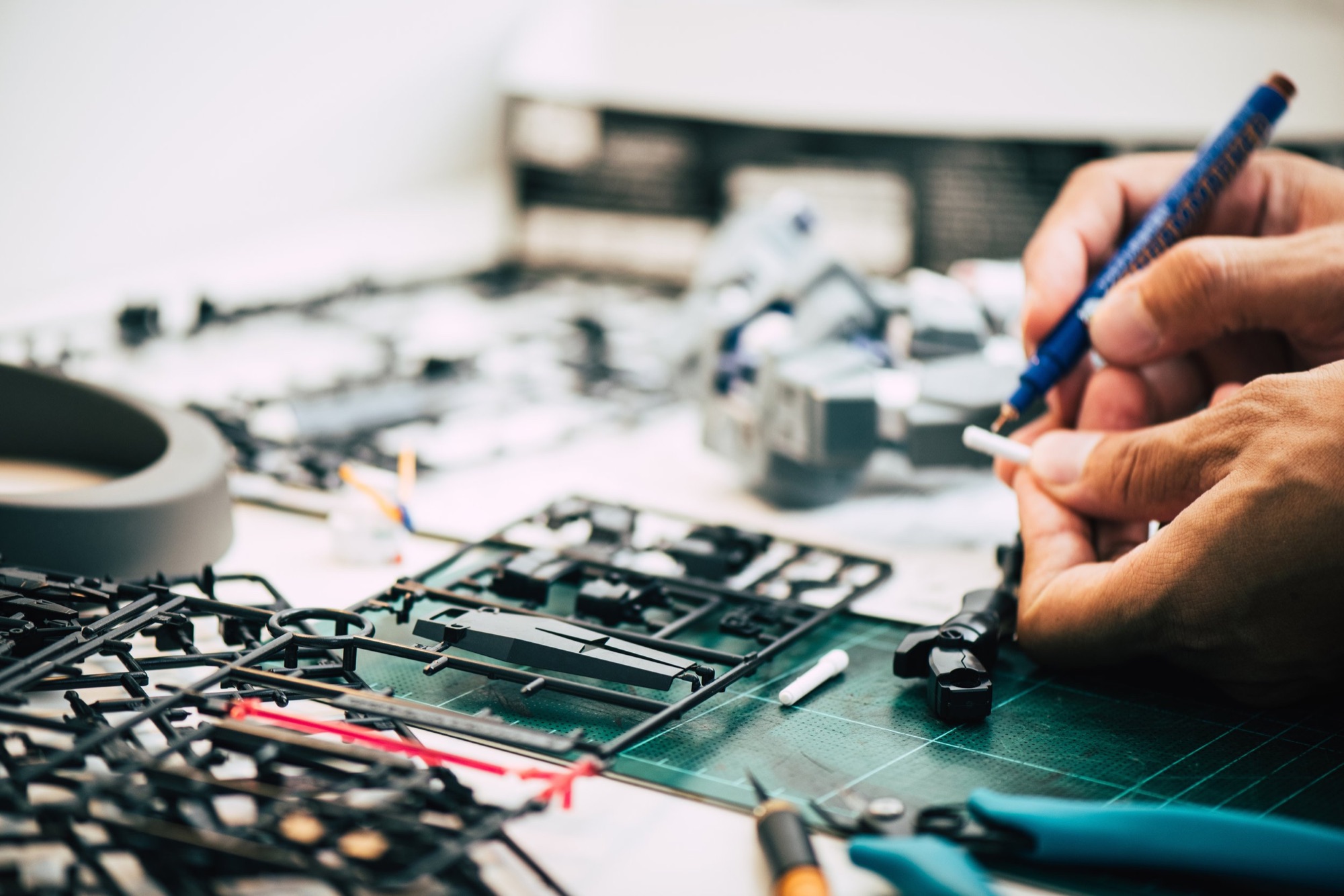 Hands assembling a small black plastic model on a green cutting mat surrounded by tools, plastic parts, and modeling supplies on a cluttered work surface.