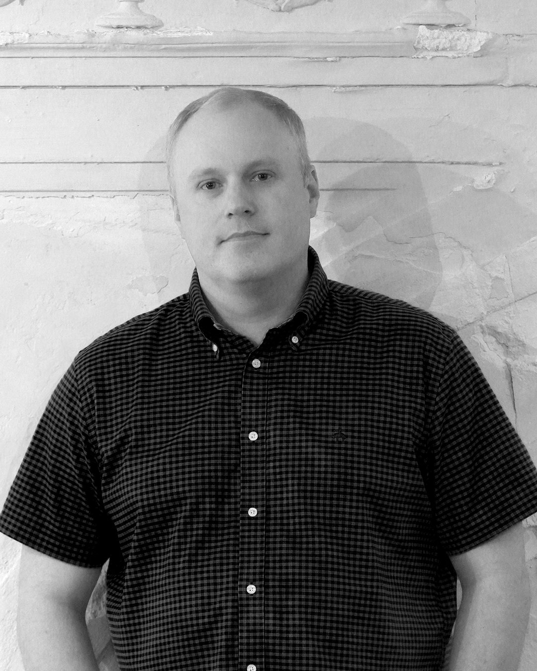 Portrait of a young man with light hair wearing a dark checked button-up shirt, standing against a textured wall background in black and white.