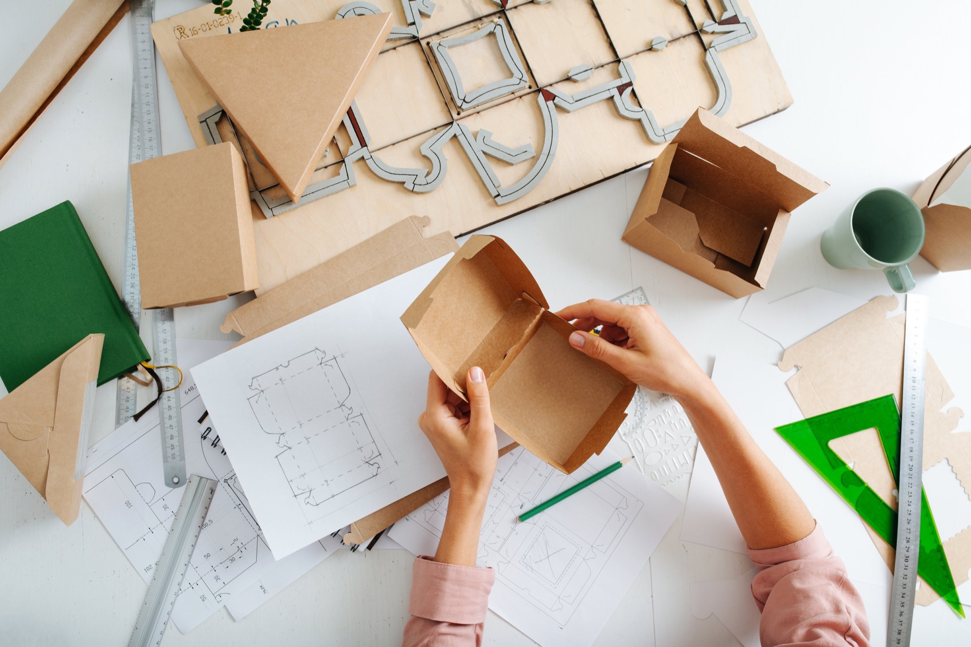A person assembling a cardboard box at a cluttered workspace with technical drawings, rulers, and other packaging materials around.